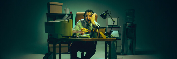Frustrated man rests head on hand, holding sandwich at desk buried in files, expressing burnout,...