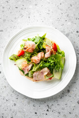 Fresh salad with crispy bacon, lettuce, cucumber, and cherry tomatoes on a white plate, placed on a speckled gray countertop. The vibrant colors and textures make it visually appealing and appetizing