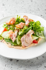 Fresh salad with crispy bacon, lettuce, cucumber, and cherry tomatoes on a white plate, placed on a speckled gray countertop. The vibrant colors and textures make it visually appealing and appetizing