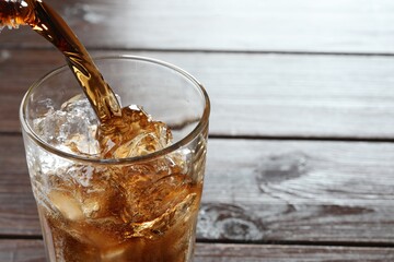 Pouring refreshing cola into glass with ice at wooden table, closeup. Space for text