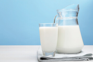 Fresh milk in glass and jug on white wooden table against light blue background. Space for text