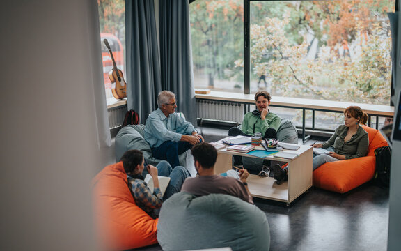 A professor conducts a discussion with students in a relaxed setting. They are seated comfortably on bean bags, fostering an informal learning atmosphere with natural light coming from large windows.