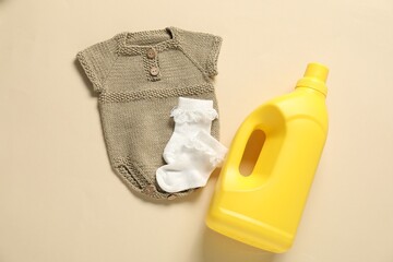 Bottle of fabric softener and baby clothes on beige background, flat lay