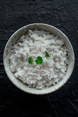 White rice, cooked. A bowl of boiled long grain rice, a simple side dish, with fresh parsley leaves, on a black slate background, top shot