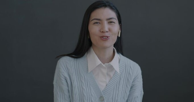 Portrait of excited young Asian woman making online video call talking waving hand using earphones on gray color background. People and communication concept.