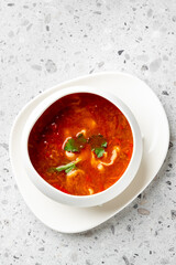 A bowl of hot, spicy soup garnished with fresh cilantro and green herbs, served on a white plate against a speckled gray background. 