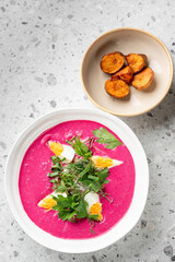 A bowl of vibrant pink beet soup with boiled egg quarters, fresh parsley, and dill, served alongside roasted potato slices on a speckled gray countertop.