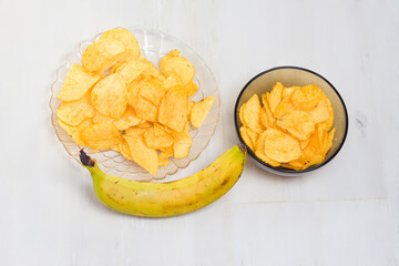 Crispy golden banana chips are served on a clear plate and in a small dark bowl alongside a fresh, ripe banana, all arranged on a white wooden table.