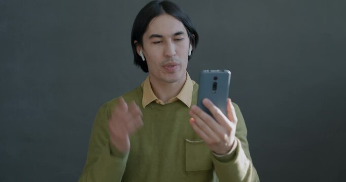 Cheerful Asian man making online video call with smartphone and earphones talking gesturing on gray color background.