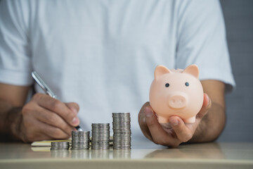 Person Holding Piggy Bank While Recording Savings, Stacks of Coins Visible on Table, Financial...