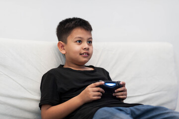 Asian young boy enjoys playing video games on couch, focused and engaged. His expression shows...