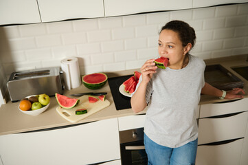 A woman casually stands in her kitchen, eating a slice of fresh watermelon. The scene radiates...