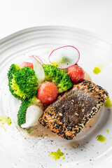 A gourmet dish featuring sesame-crusted fish, fresh broccoli, radish slices, and cherry tomatoes, elegantly plated with sauce and green oil droplets on a white plate.
