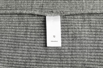 Clothing label with size and material content on grey garment as background, top view