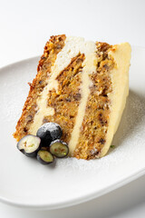 A slice of carrot cake with cream cheese frosting on a white plate, garnished with powdered sugar and fresh blueberries. Close-up view of a delicious homemade dessert.