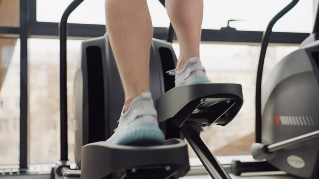 Close up of gym enthusiast wearing sneakers powering elliptical trainer pedal with controlled motion during dynamic cardio workout in bright sleek modern fitness center by large windows