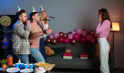 Young adult birthday party, men and woman friends in festive hats meeting and surprising birthday girl, blowing in horns and laughing, happy positive expression.