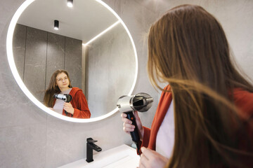 Young woman in a red robe using a hairdryer to style her long hair in a sleek bathroom with soft...
