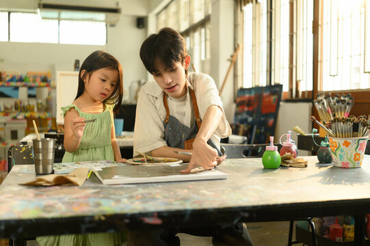 A young LGBT teacher explains painting steps to a little girl in an art class surrounded by brushes, paints, and creative tools in a bright and inspiring studio