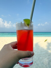 Female hand holding glass with red cocktail against ocean in Maldives. Tropical resort vacation