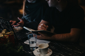 Two people engage in a discussion, working on notes and a laptop at a cafe table. The environment is cozy with a festive atmosphere, featuring a warm ambiance and gift-wrapped presents.