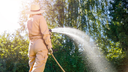 Woman in gardening overalls with garden hose with visible stream of water flowing and garden...