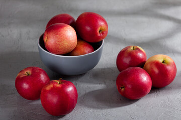 A gray ceramic bowl filled with red apples sits on a textured gray surface, with several apples scattered around it. Soft natural light and shadows create a calm, minimalist food scene.