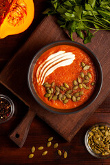 A dish bright pumpkin soup garnished with cream and pumpkin seeds on a wooden chopping board, fresh parsley, peppercorns and half a pumpkin for decoration. Homegrown organic eco food 