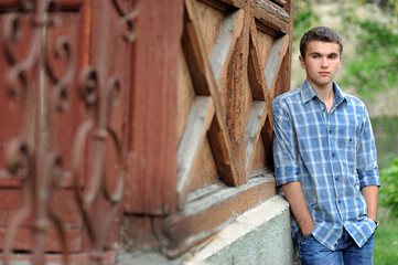 Portrait of a young man in a checkered shirt, hands in pockets, standing near a carved porch, retro...