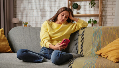 Obraz premium Young woman spending day at home sitting on the sofa in the living room, holding smartphone, watching social media, bored expression.