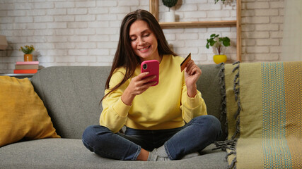 Fototapeta premium Young woman spending day at home sitting on the sofa in the living room, typing card number on smartphone, shopping buying online, positive expression.