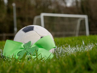 Green bow with Irish shamrock by a classic football or soccer ball on a green grass training field. Goal post in the background. Supporting national team of Ireland concept.