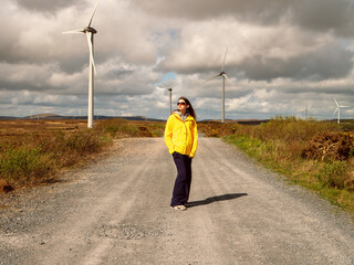 Young teenager girl in yellow jacket standing on a road, Wind turbines and vast wild fields in the...