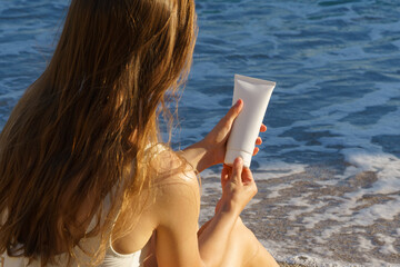 Young woman with flowing hair opens a white mockup tube of cream on the background of sea water on the beach. Concept of sun protection products, care cosmetics, beauty and aesthetics.