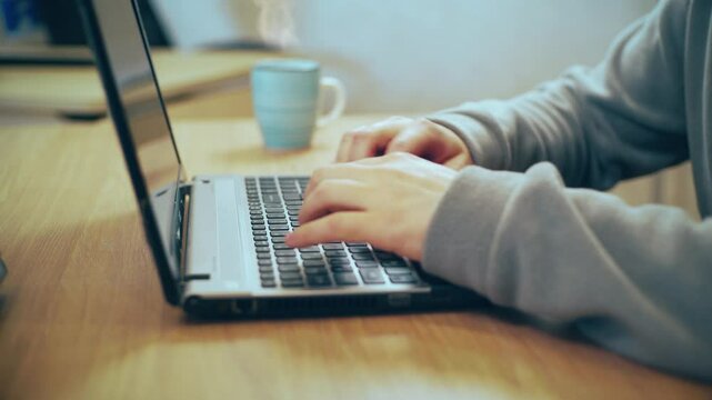 close-up of a man's hands using a computer keyboard, laptop, to find what they are interested in, with a steaming coffee mug in the background.