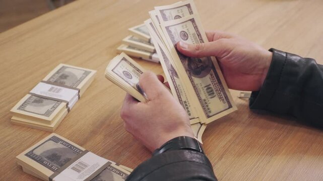 close-up of male hands counting a stack of 100 dollar bills, with stacks of dollars in the background.