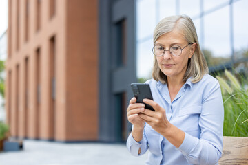 A mature woman with glasses, sitting outdoors, focused on her smartphone. She is engaged in messaging or browsing on her mobile device.