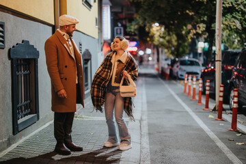 A couple dressed in warm clothing shares a lighthearted moment during an evening stroll in the city. The setting is urban, with streetlights illuminating the path and cars parked along the street.