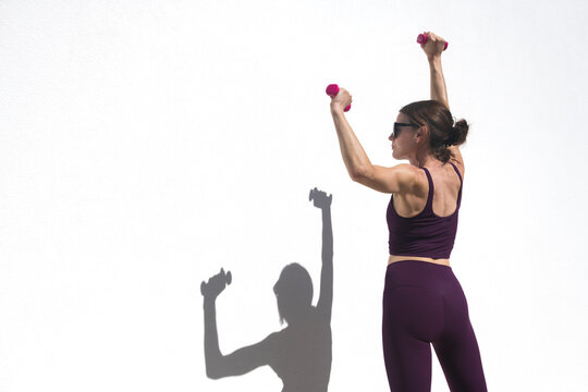 Athletic woman exercising with hand weights doing arm exercises outdoors by a white wall