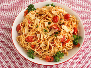Overhead view of bowl of spaghettini with cherry tomatoes and garnished with basil, parsley and shaved parmesan against a red background