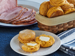Two sweet potato biscuits, one open with butter on it, in front of a basket of more biscuits and a sliced ham in the background