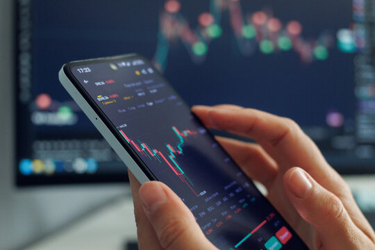 A person's hands hold a smartphone displaying a stock market chart with red and green candles, indicating price fluctuations, against a blurred background showing more financial data 