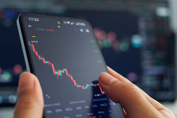 A person's hands hold a smartphone displaying a declining stock market chart with mostly red candlesticks, indicating a negative trend, against a blurred backdrop of a larger financial data screen.