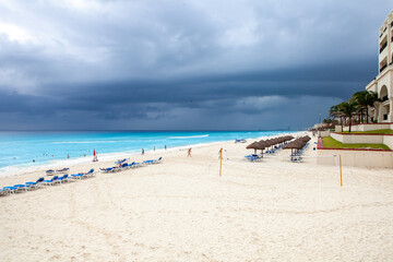 Vista ampla da praia de Cancún com mar azul-turquesa, espreguiçadeiras vazias e céu nublado sobre o litoral caribenho.