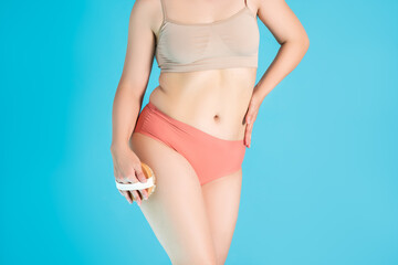 Slim woman in beige top and pink panties with dry massage brush on blue background, body care...