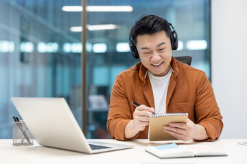 A smiling asian man wearing headphones takes notes while looking at a laptop in an office...