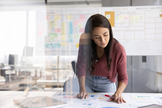 Developing project. Busy young woman business professional data analyst bend over desk with paper documents review reports read check statistic information. View through glass office wall. Copy space