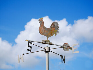A rusty cockertal weathervane against a summer sky of white fluffy clouds showing the points of the compass