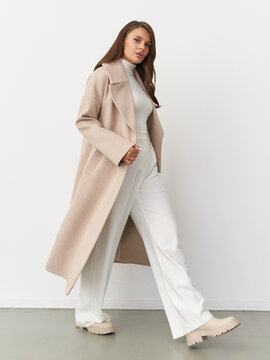 Stylish woman in beige coat, white turtleneck, and trousers walks confidently. Light brown hair, casual yet sophisticated look. Neutral tones enhance the elegant and modern outfit
