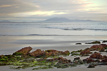 Red rocks covered by green moss on the beach sand,  waves breaking on water edge, foggy morning with cloudy sky in Itacuruca beach, Cananeia, Sao Paulo state